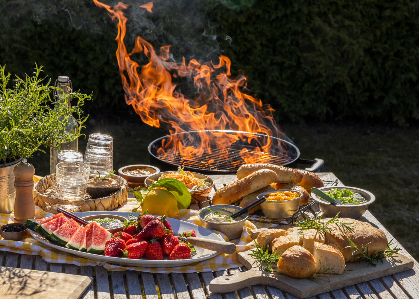 Et sommermåltid ute med grill i flammer, brød, frukt, salater og tilbehør på bordet