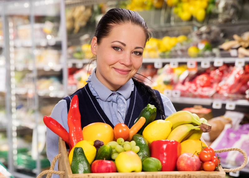 Smilende butikkmedarbeider holder en kurv fylt med friske grønnsaker og frukt i en dagligvarebutikk