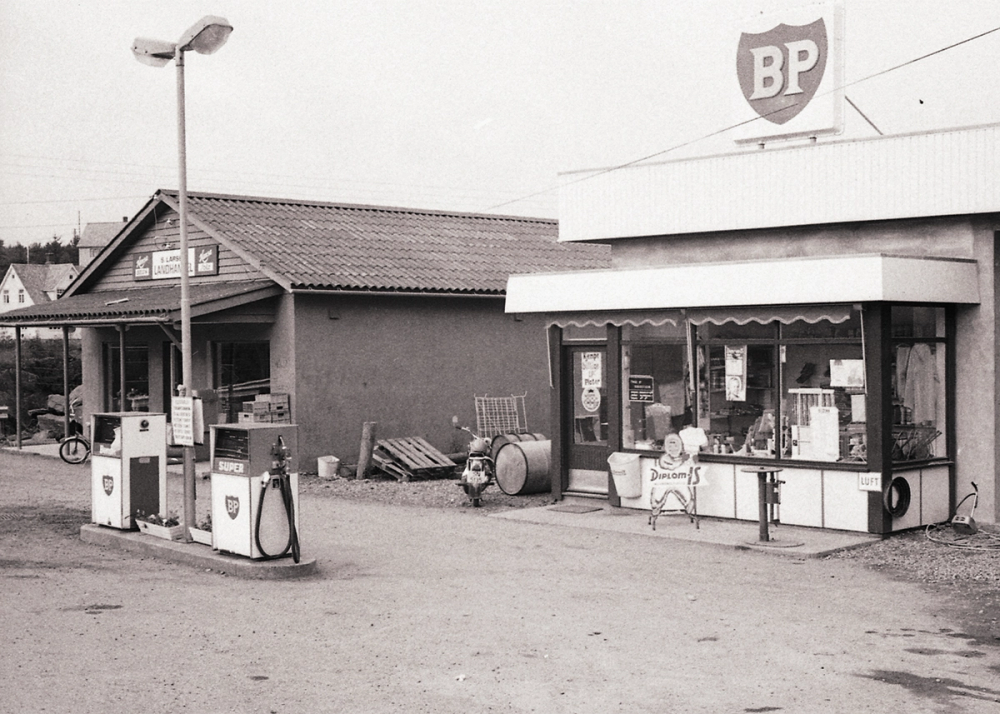 Svart-hvitt-bilde av en bensinstasjon med BP-logo, gamle bensinpumper og en liten butikk med skilt for Diplomis