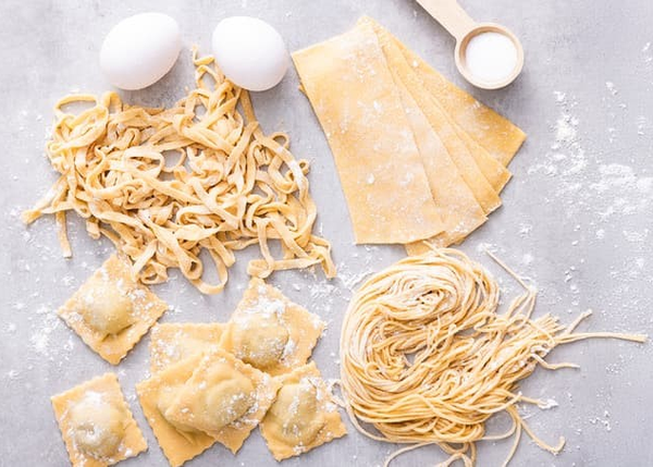 Ulike typer fersk, hjemmelaget pasta – tagliatelle, ravioli, lasagneplater og spagetti – lagt på et bord med mel og egg
