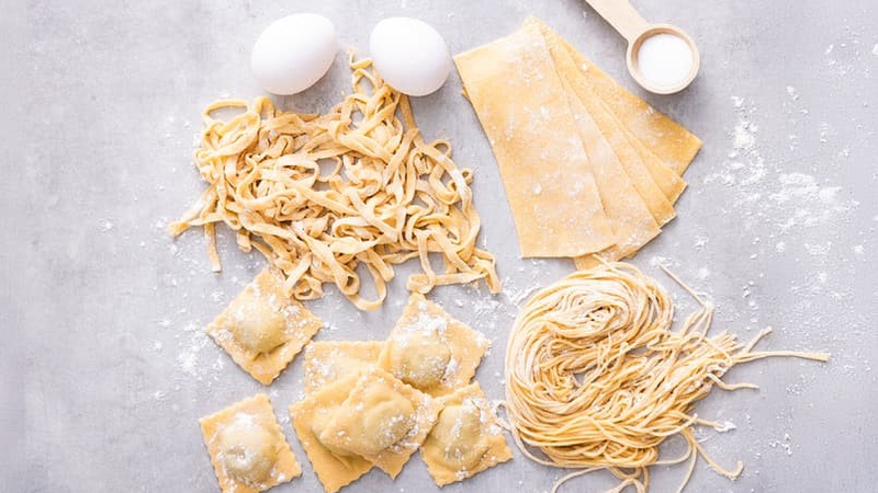 Ulike typer fersk, hjemmelaget pasta – tagliatelle, ravioli, lasagneplater og spagetti – lagt på et bord med mel og egg
