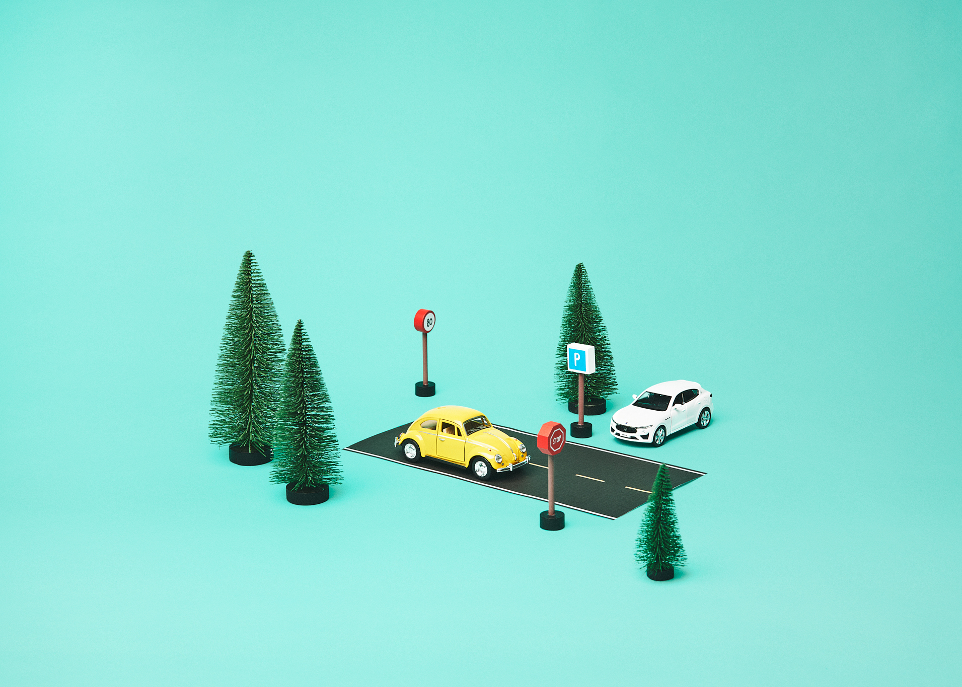 Modellbiler og trafikkskilt på en liten vei omgitt av trær mot turkis bakgrunn