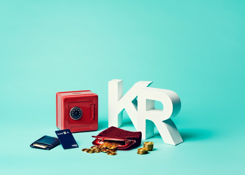 Rød safe, lommebøker, bankkort og mynter foran et stort hvit KR-symbol på blå bakgrunn