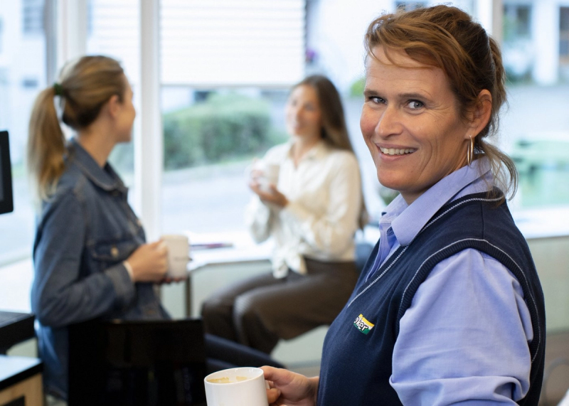 En Joker-ansatt smiler til kamera, to kunder sitter og prater i bakgrunnen