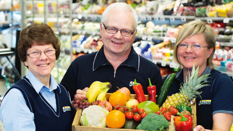 Tre butikkansatte smiler og holder en kasse fylt med fersk frukt og grønnsaker i dagligvarebutikk
