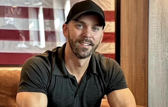 Man in a casual black cap and dark polo shirt sitting indoors, smiling warmly, with an American flag displayed in the background.