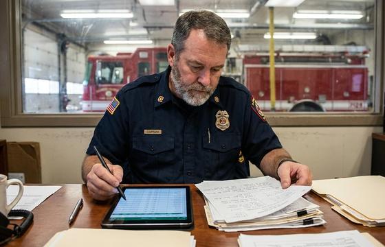 Fire captain cross-checking a tablet data record against a printed incident report at a station desk