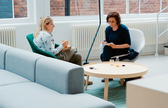 2 women sit in a modern office lounge having a focused conversation with notebooks and drinks on a round wooden table.