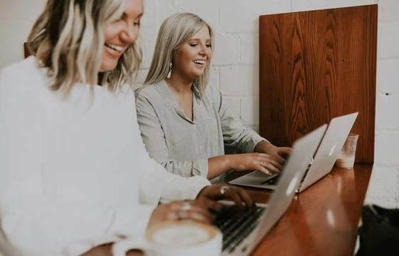 Employee engagement of 2 women working on laptops at a wooden table in a cozy cafe.