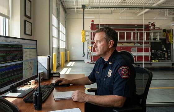 Fire company officer reviewing fire department operations data at a station watch desk, fire engine in the apparatus bay behind