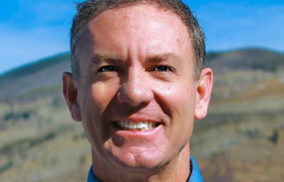 Close-up of a smiling man outdoors with a mountainous landscape in the background, under a bright blue sky, conveying positivity and natural beauty