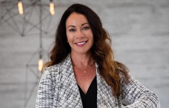 The image shows a woman with long, wavy brown hair, wearing a black top under a textured, light-colored blazer. She has a warm smile, and her background is a modern, industrial-style setting with a concrete wall and hanging lights, adding a professional yet approachable feel to the scene.