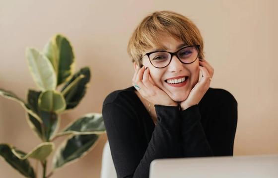 Smiling woman in glasses sitting at a desk with a plant in the background, representing a positive workplace environment and engagement.