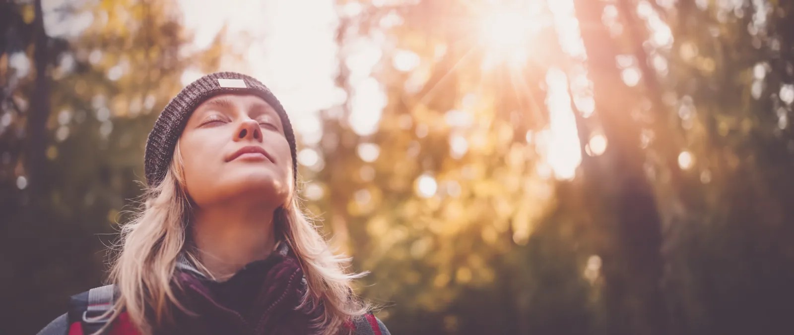 Woman in a beanie standing in a sunlit forest with her eyes closed