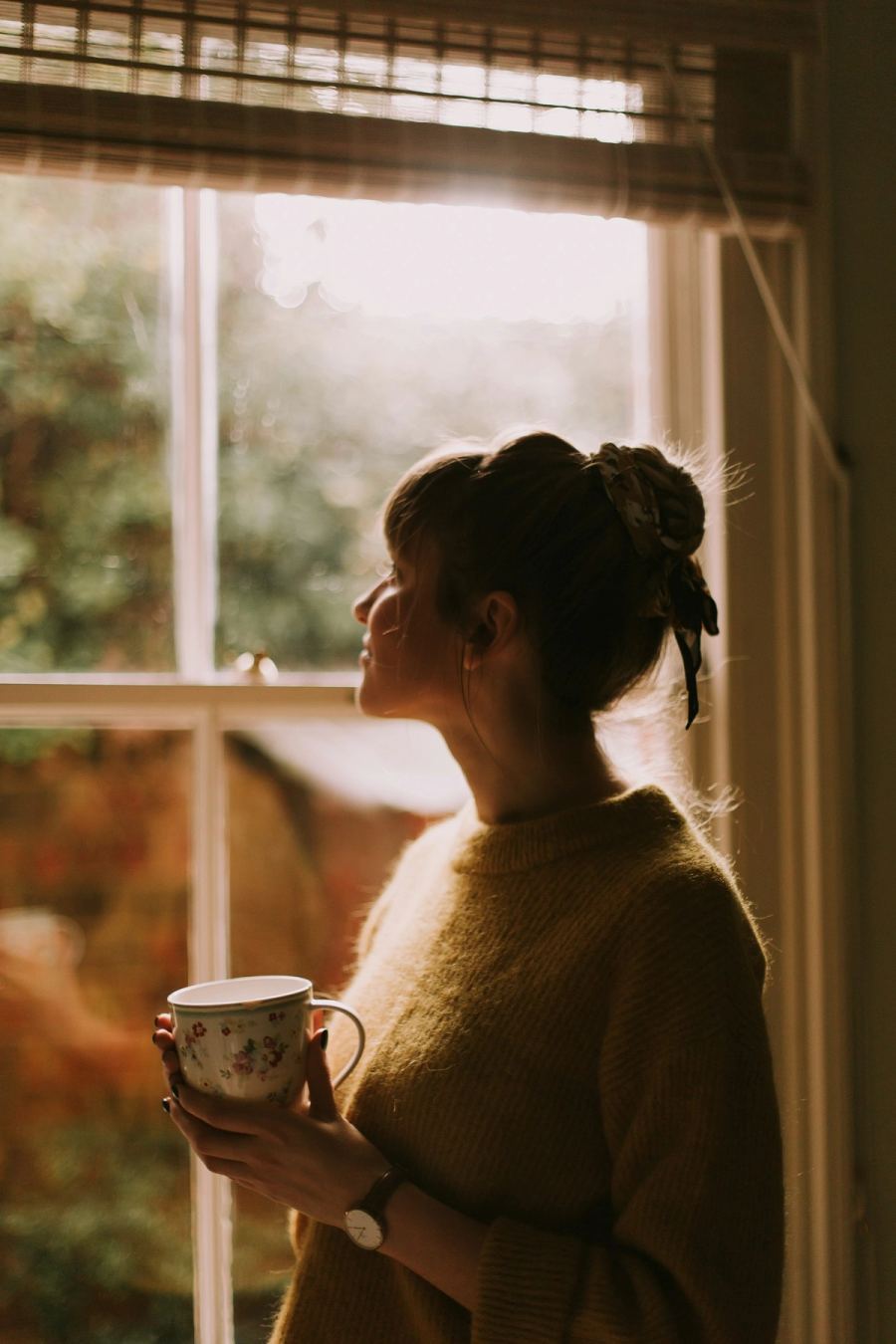 Woman in a cosy sweater holding a cup of tea beside a sunlit window in autumn