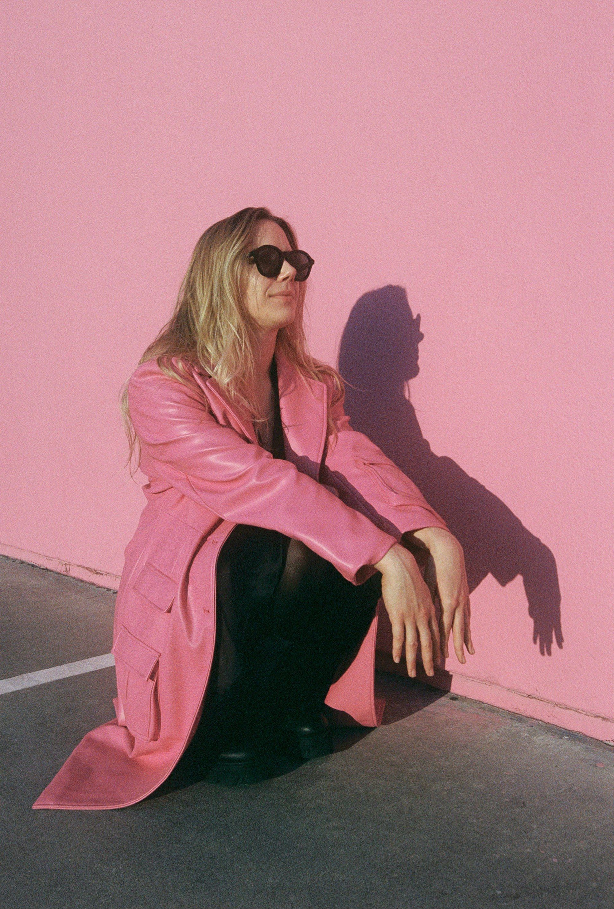 Woman in a pink coat crouching against a pink wall