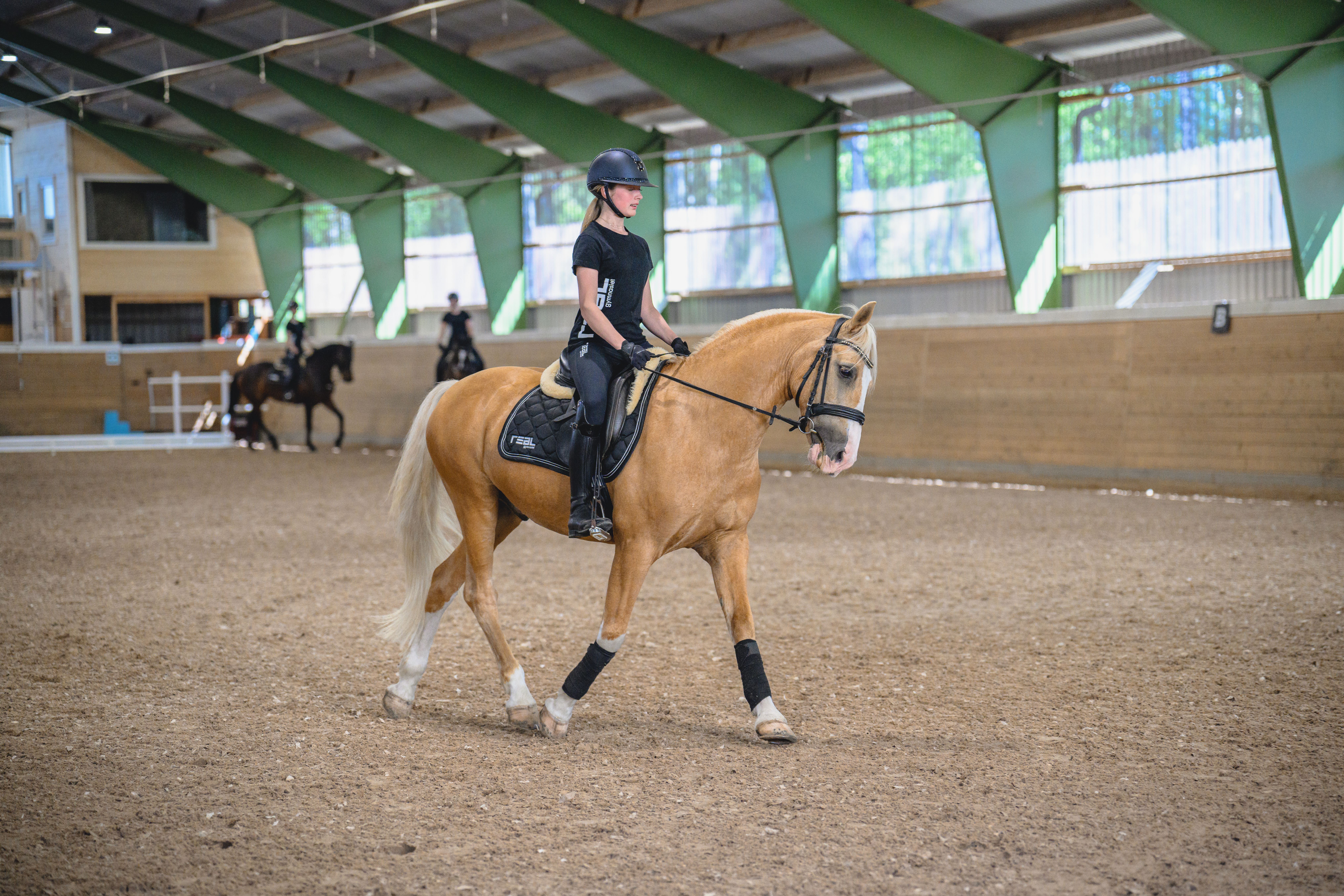 En ung ryttare med ridhjälm och svart jacka står bredvid en brun häst i en inomhusridanläggning