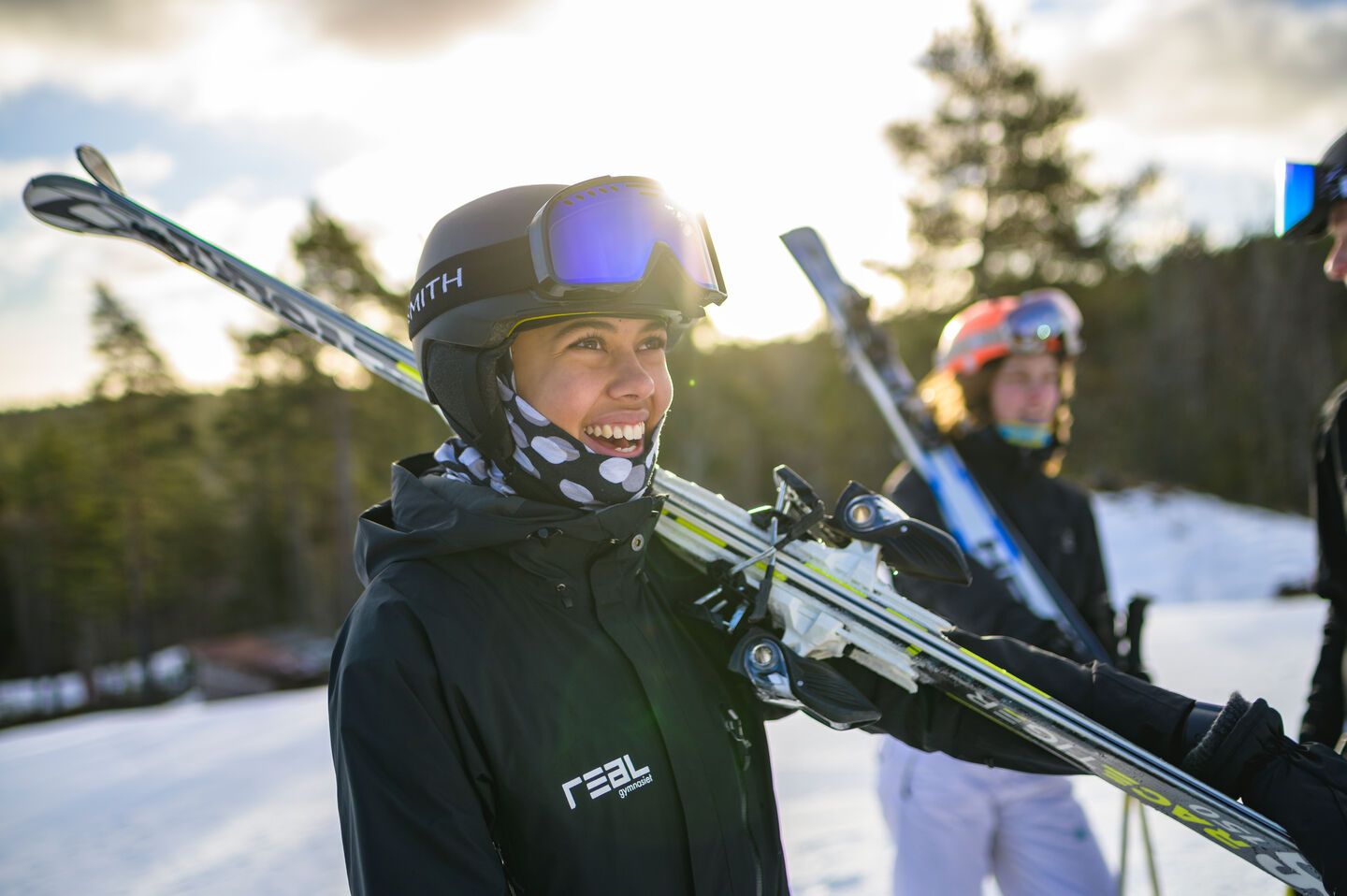 En glad elev med hjälm och skidglasögon bär sina skidor över axeln i solsken på fjället.