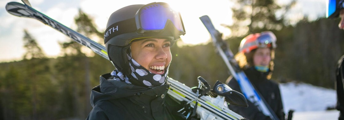 En glad elev med hjälm och skidglasögon bär sina skidor över axeln i solsken på fjället.