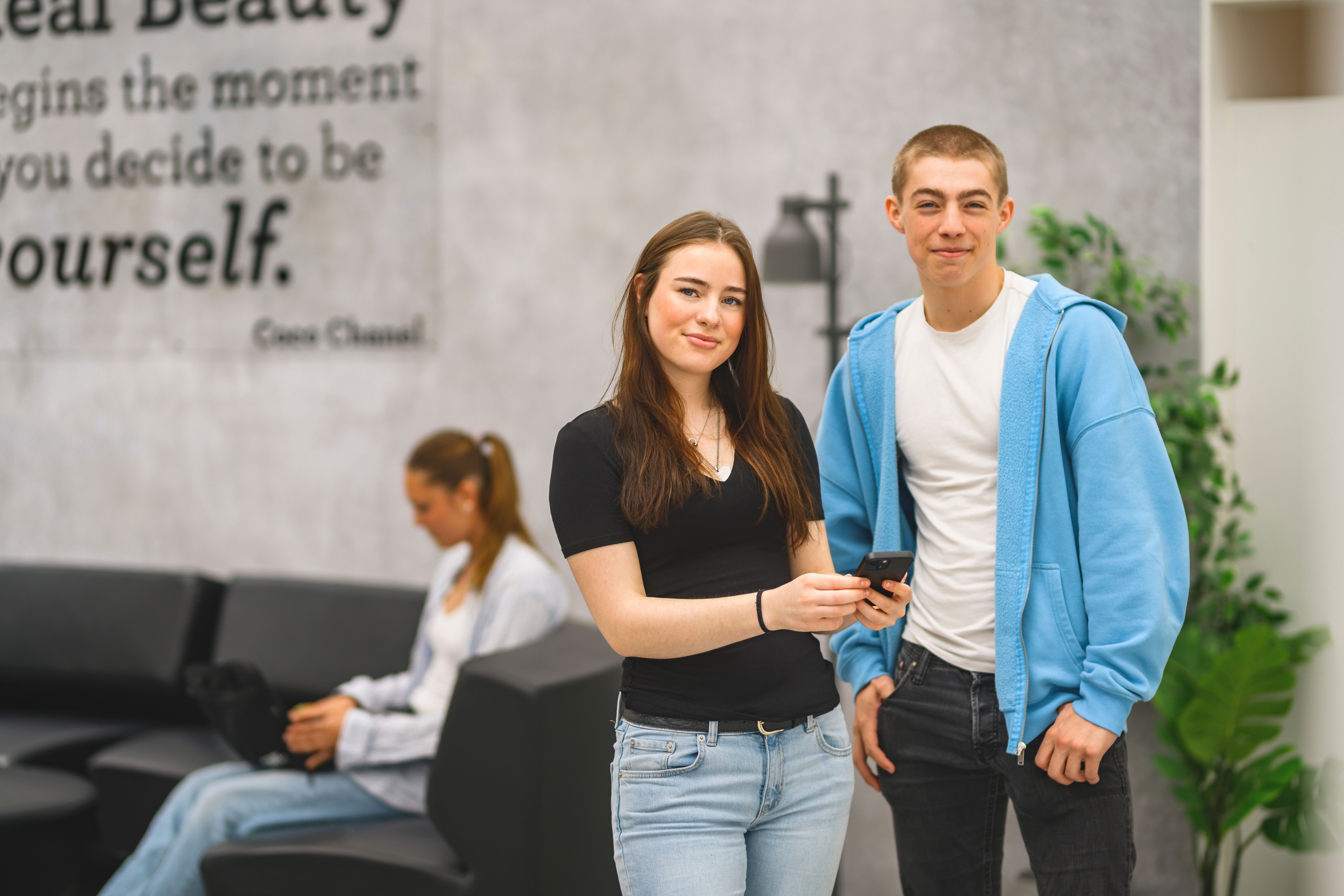 Två elever står bredvid varandra i ett uppehållsrum på skolan och ler glatt mot kameran.