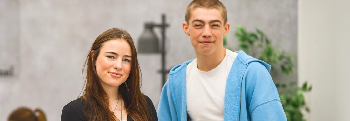Två elever står bredvid varandra i ett uppehållsrum på skolan och ler glatt mot kameran.