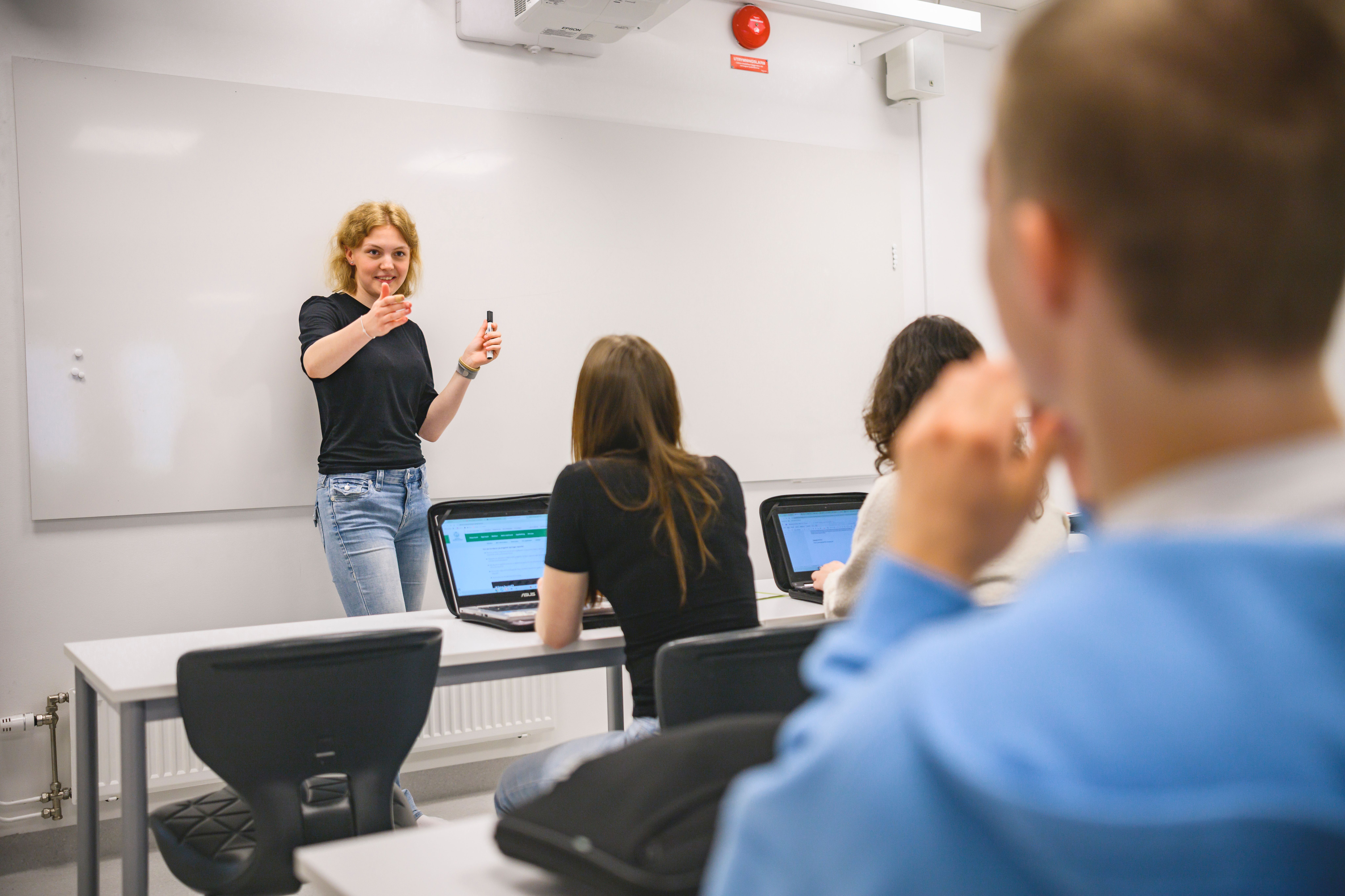 En elev står framför en whiteboard i ett klassrum och pekar på en klasskompis.