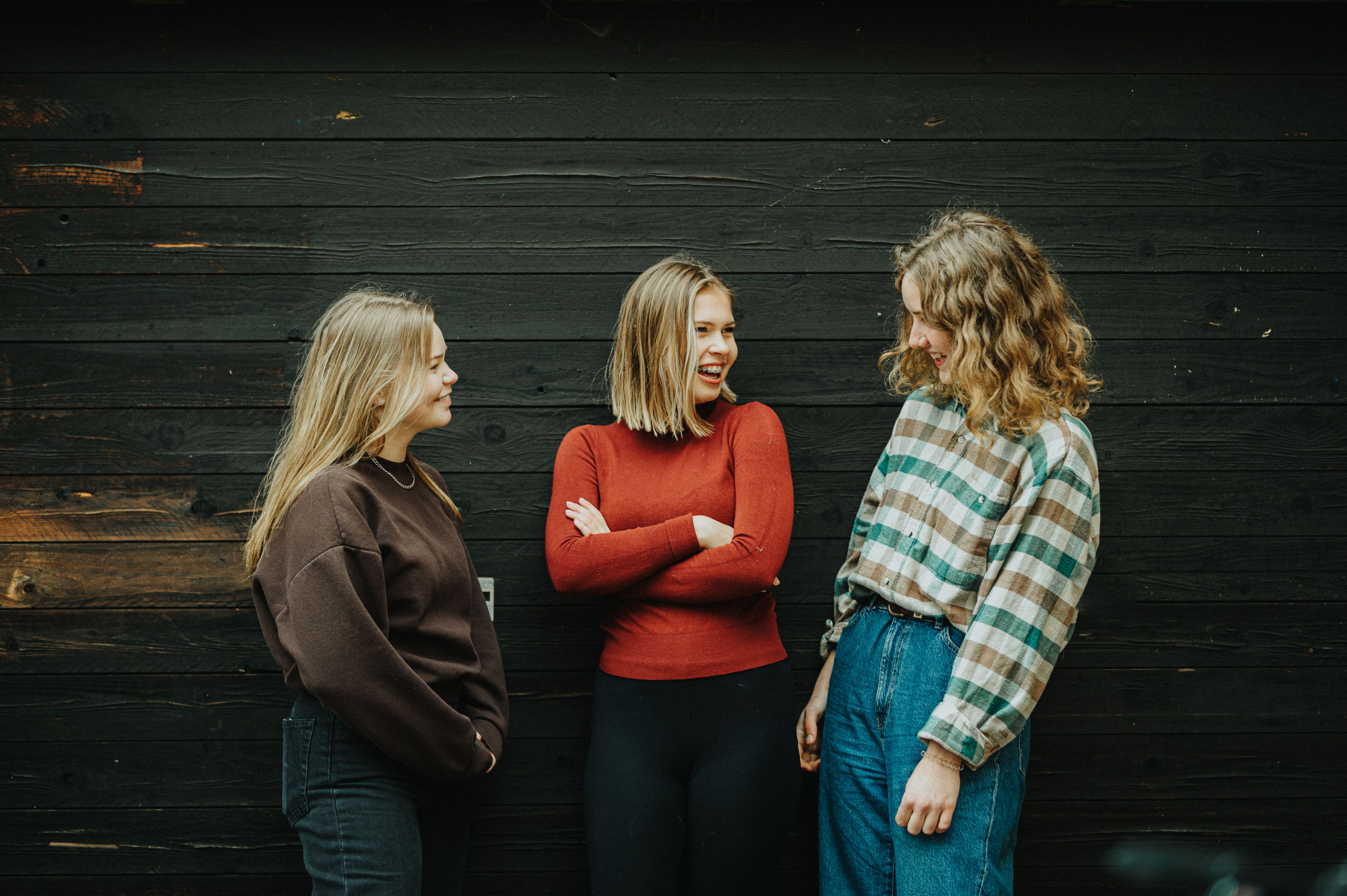 Tre tjejer står och pratar och ler vid mörk vägg