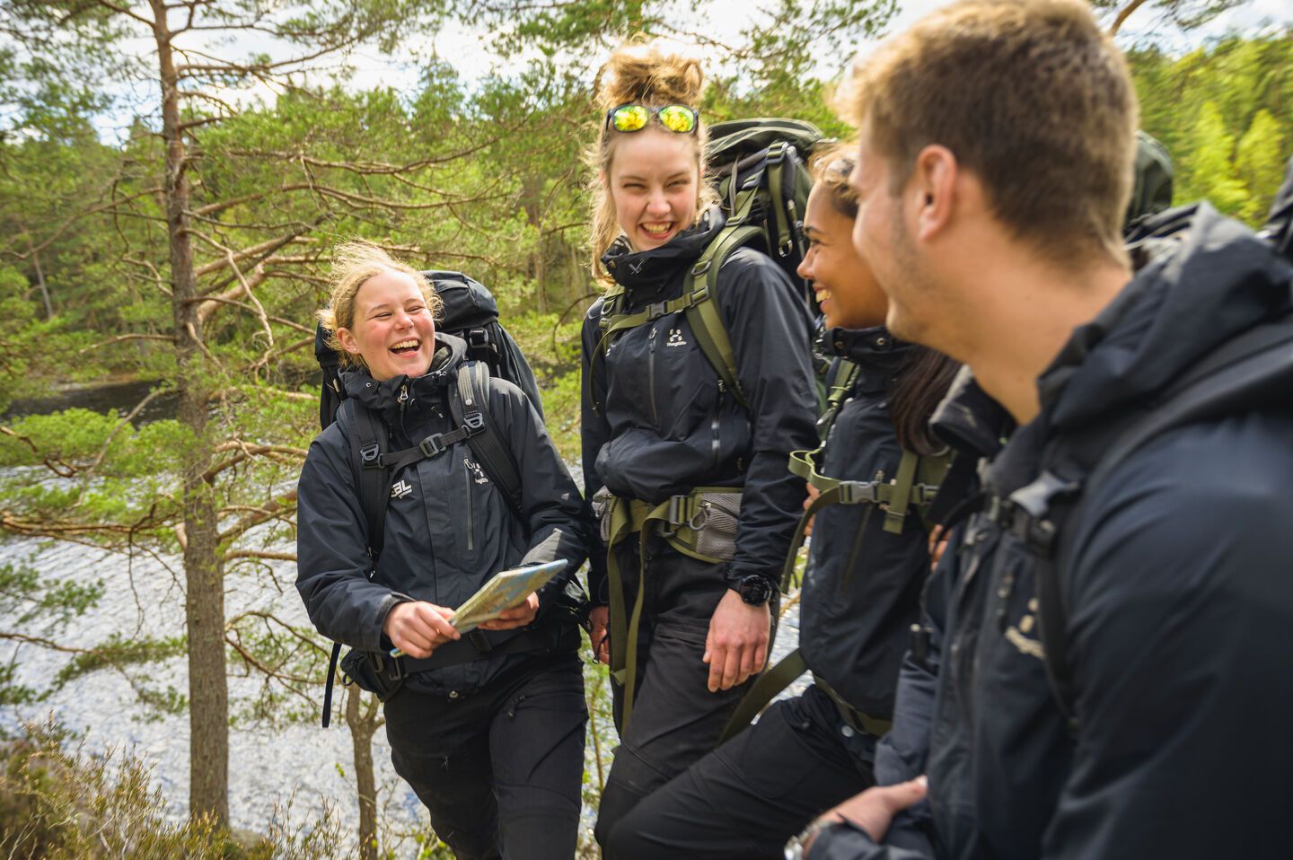 Fyra elever i svarta friluftskläder skrattar tillsammans under en vandring vid en sjö i skogen.