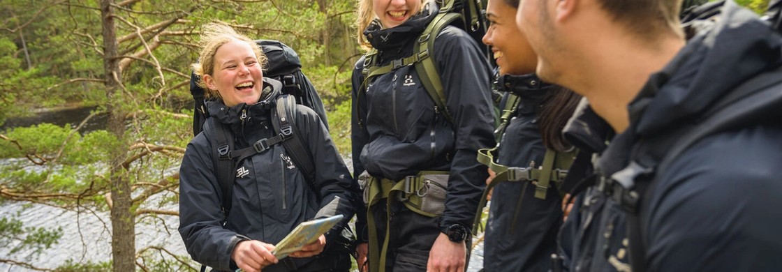 Fyra elever i svarta friluftskläder skrattar tillsammans under en vandring vid en sjö i skogen.