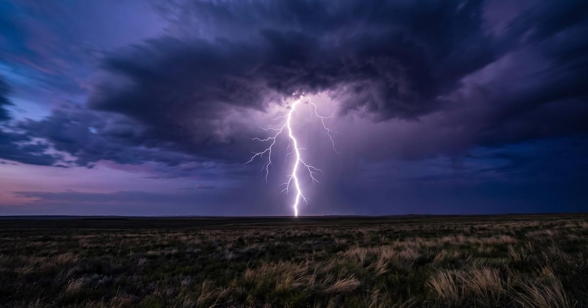 A bright lightning bolt striking from a dark storm cloud over an open field.