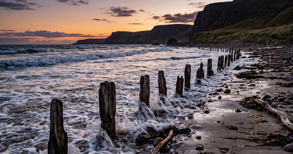 A quiet shoreline at dusk with waves eroding the sand near small wooden markers.
