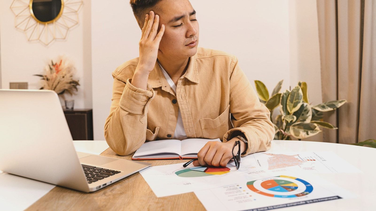 Businessman holding head in frustration, surrounded by documents and laptop at home office.