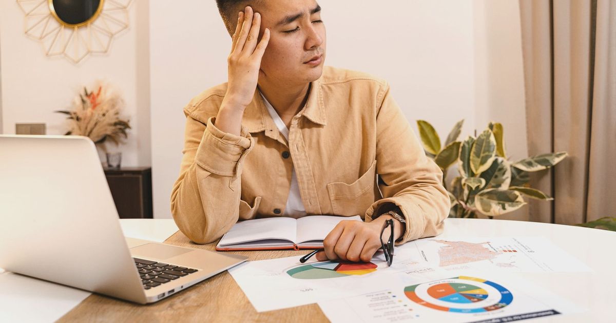 Businessman holding head in frustration, surrounded by documents and laptop at home office.