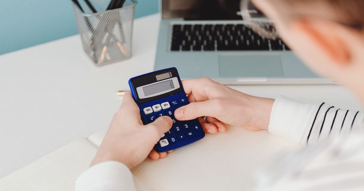 Close-up of hands using a blue calculator at a desk with a laptop and stationery.