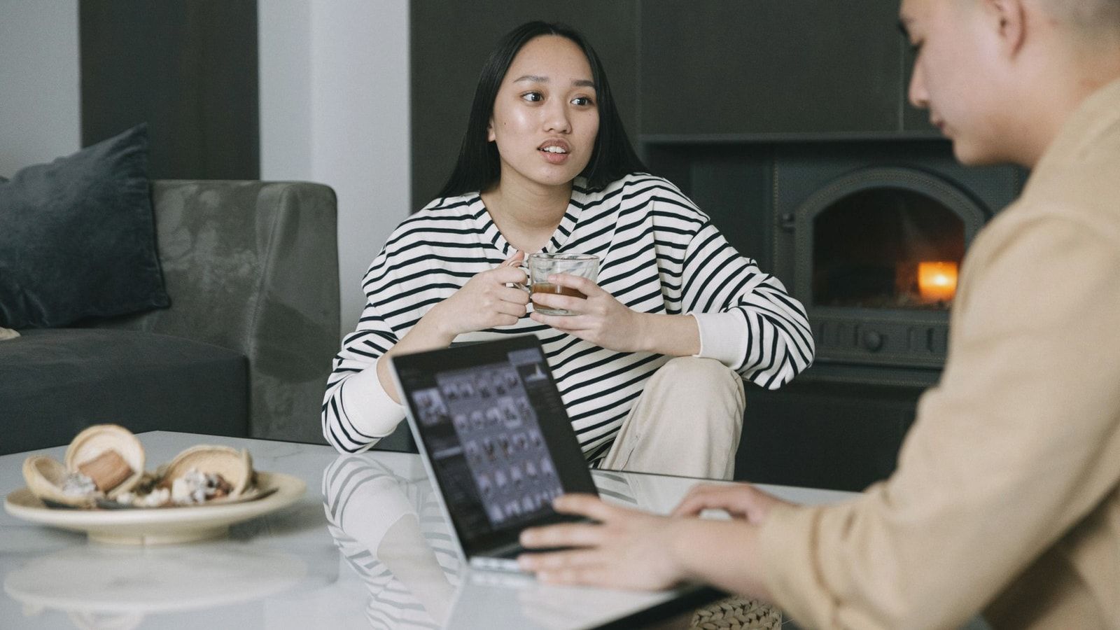 Asian couple discussing while drinking coffee and using a laptop near a cozy indoor fireplace.