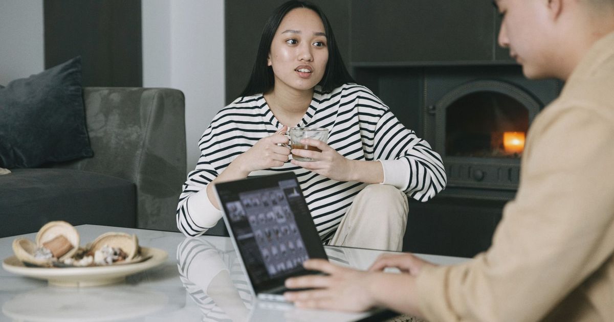 Asian couple discussing while drinking coffee and using a laptop near a cozy indoor fireplace.