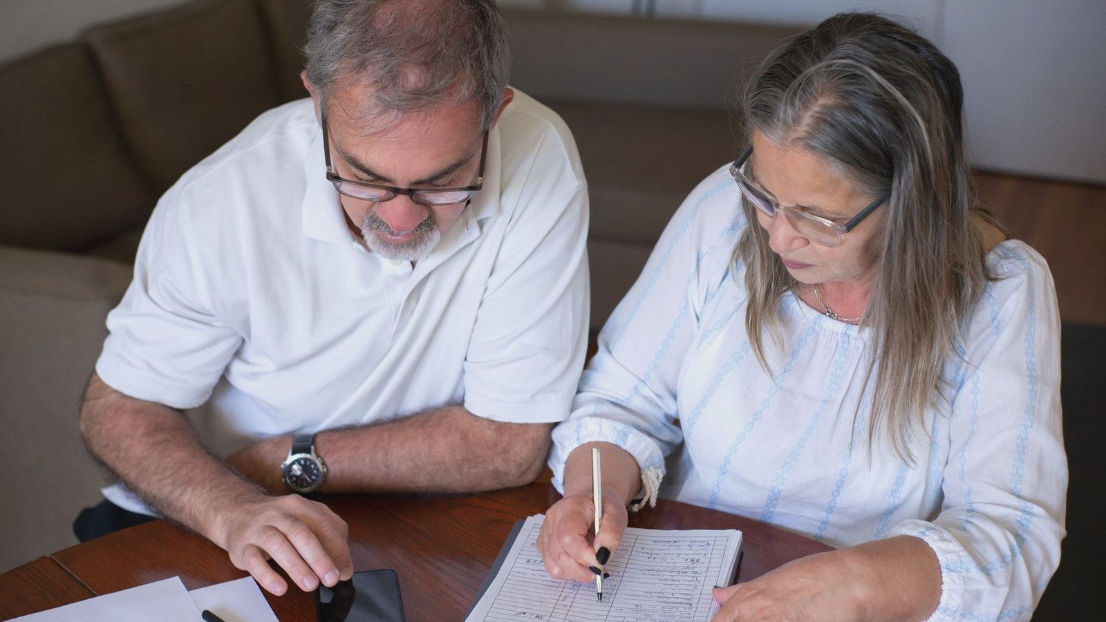 Elderly couple reviewing documents, using smartphone for online banking at home.