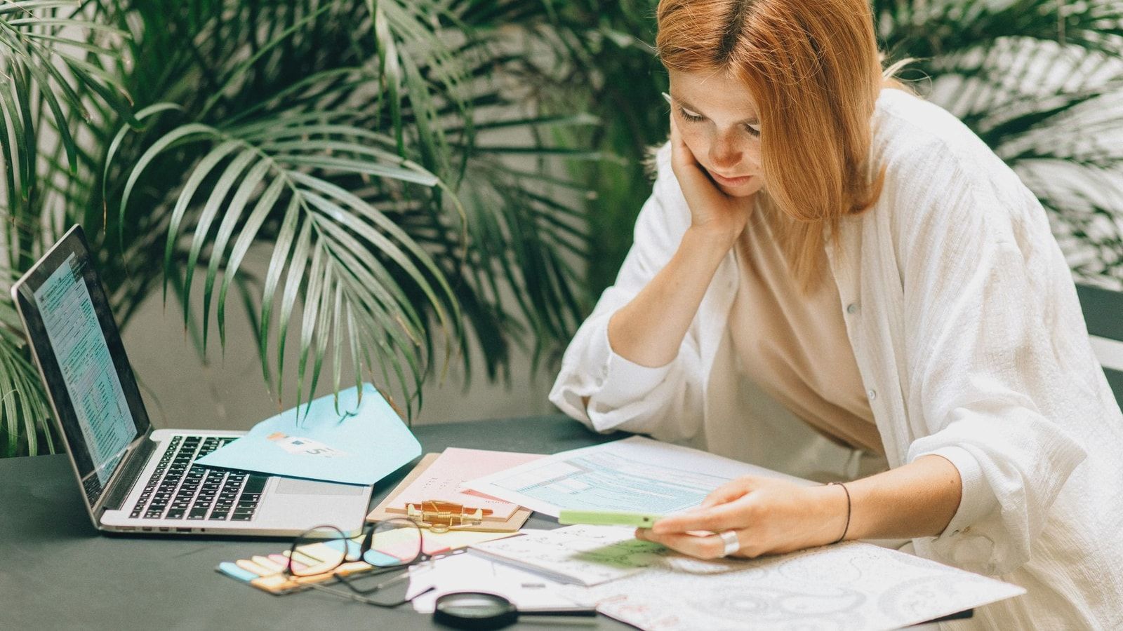 Woman in a thoughtful pose reviewing documents on a table with a laptop, surrounded by greenery.