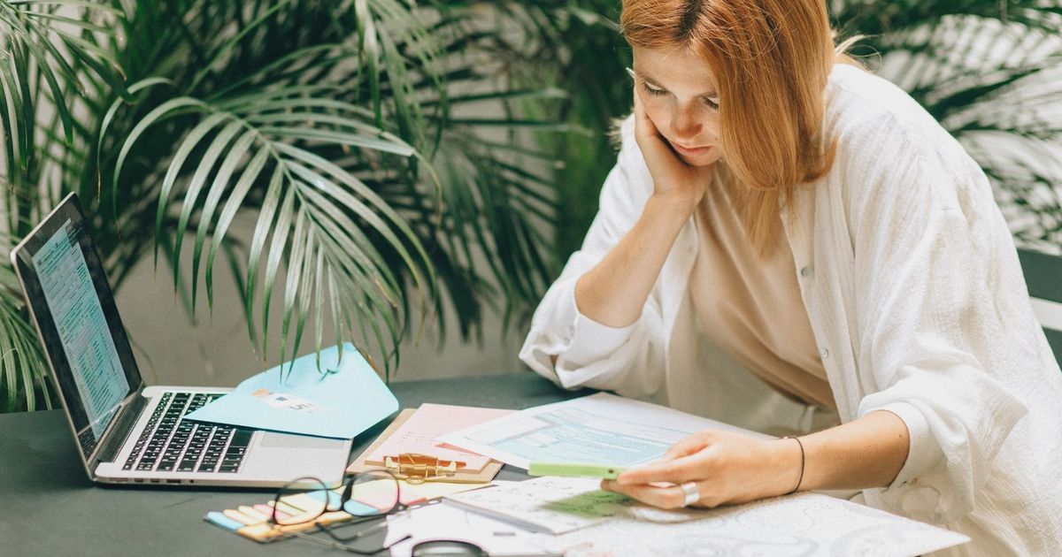 Woman in a thoughtful pose reviewing documents on a table with a laptop, surrounded by greenery.