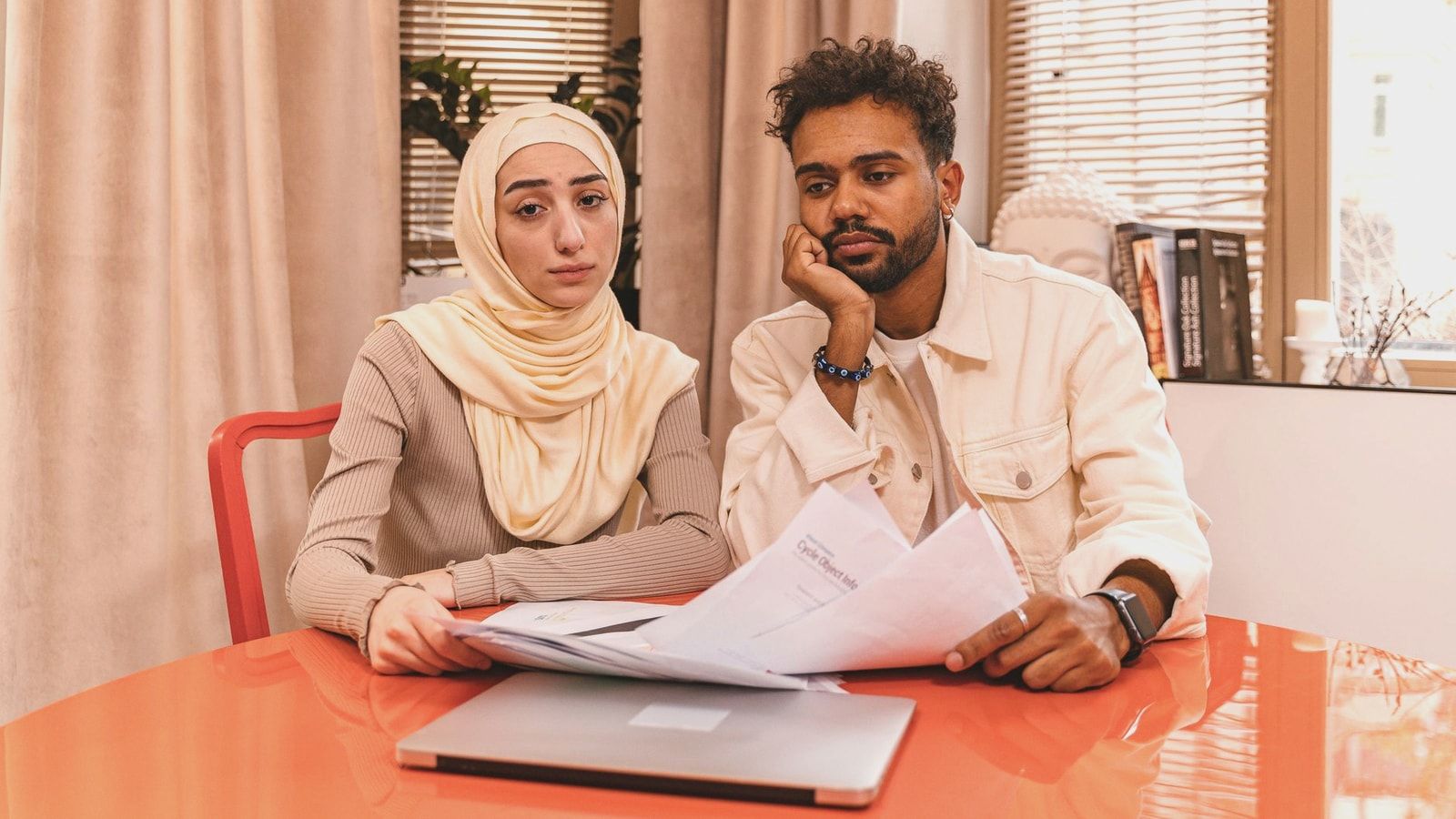 A couple in casual attire sits indoors at a table reviewing financial documents with a laptop.