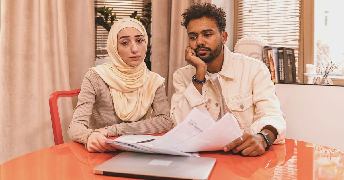 A couple in casual attire sits indoors at a table reviewing financial documents with a laptop.