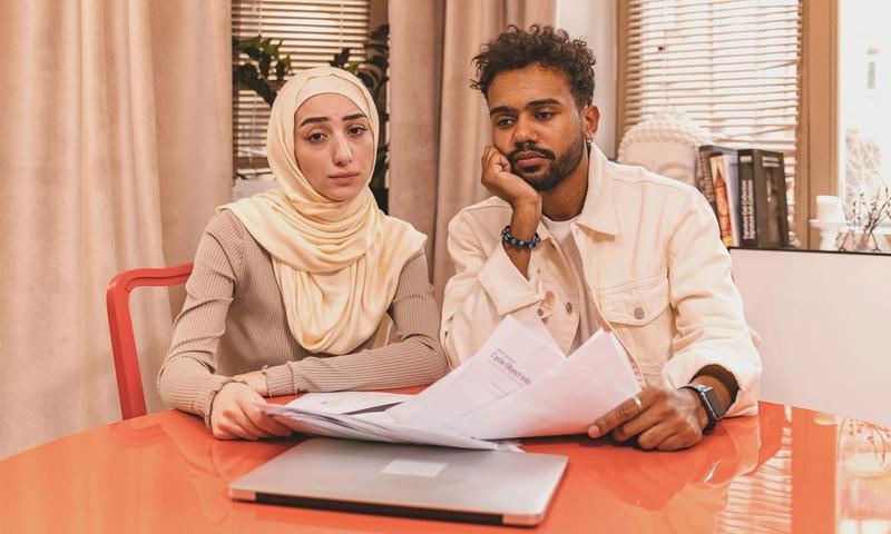 A couple in casual attire sits indoors at a table reviewing financial documents with a laptop.