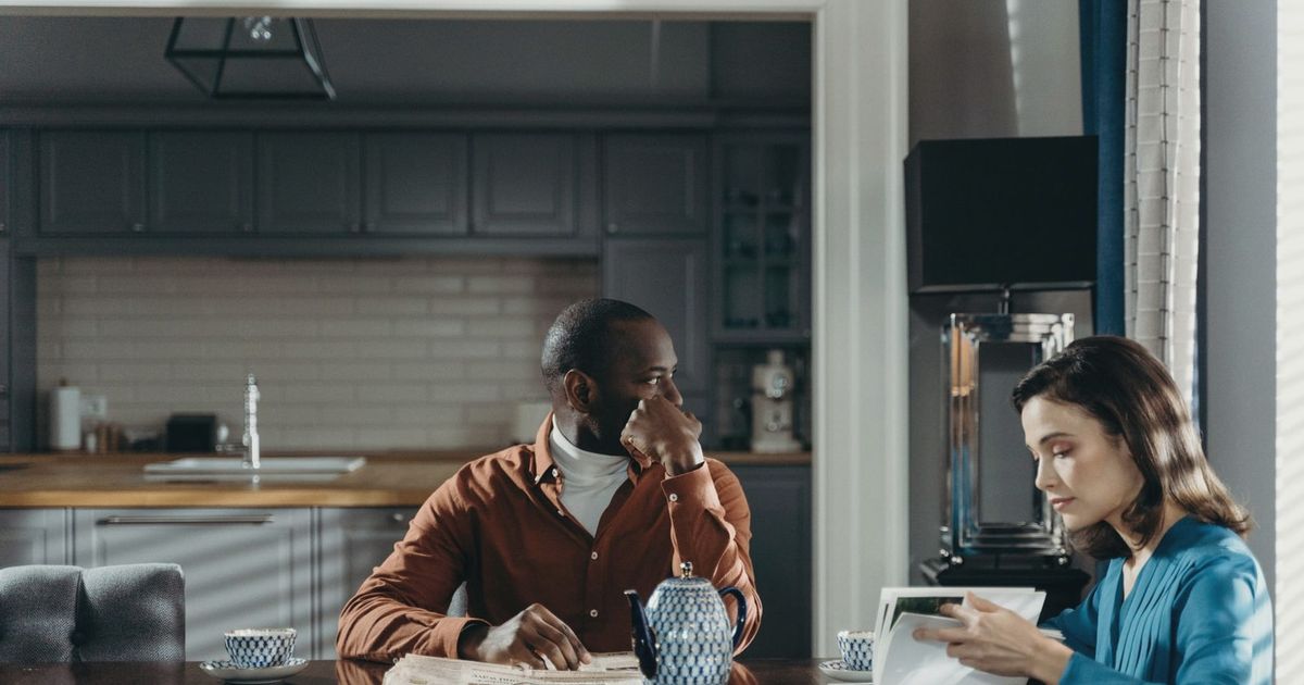A couple sits at a dining table in a modern kitchen, reading and relaxing.