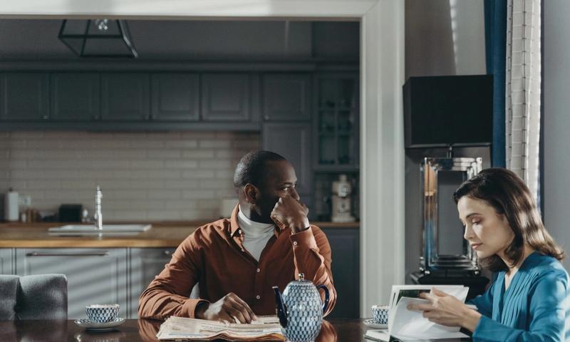 A couple sits at a dining table in a modern kitchen, reading and relaxing.