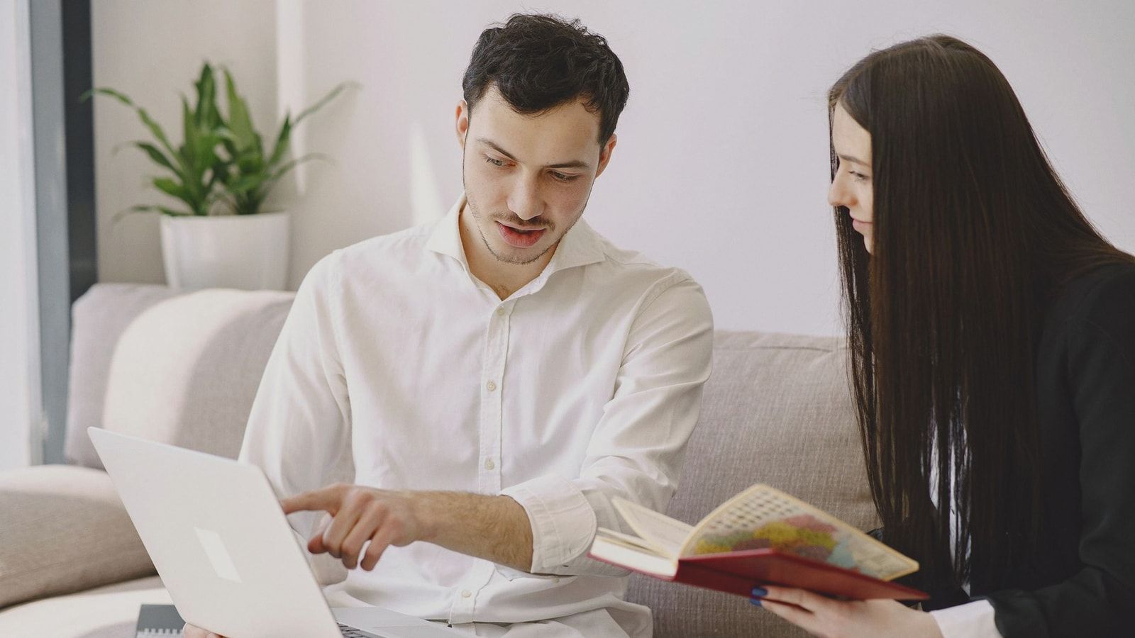 Male worker in formal wear showing with finger at laptop while discussing project with female partner with open copybook and sitting on sofa