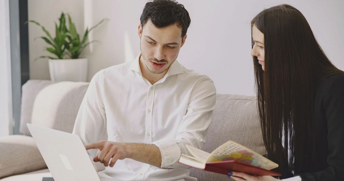 Male worker in formal wear showing with finger at laptop while discussing project with female partner with open copybook and sitting on sofa