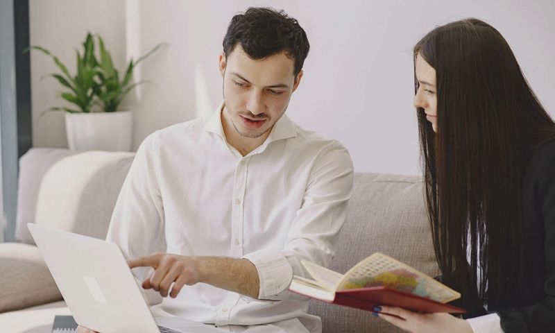 Male worker in formal wear showing with finger at laptop while discussing project with female partner with open copybook and sitting on sofa