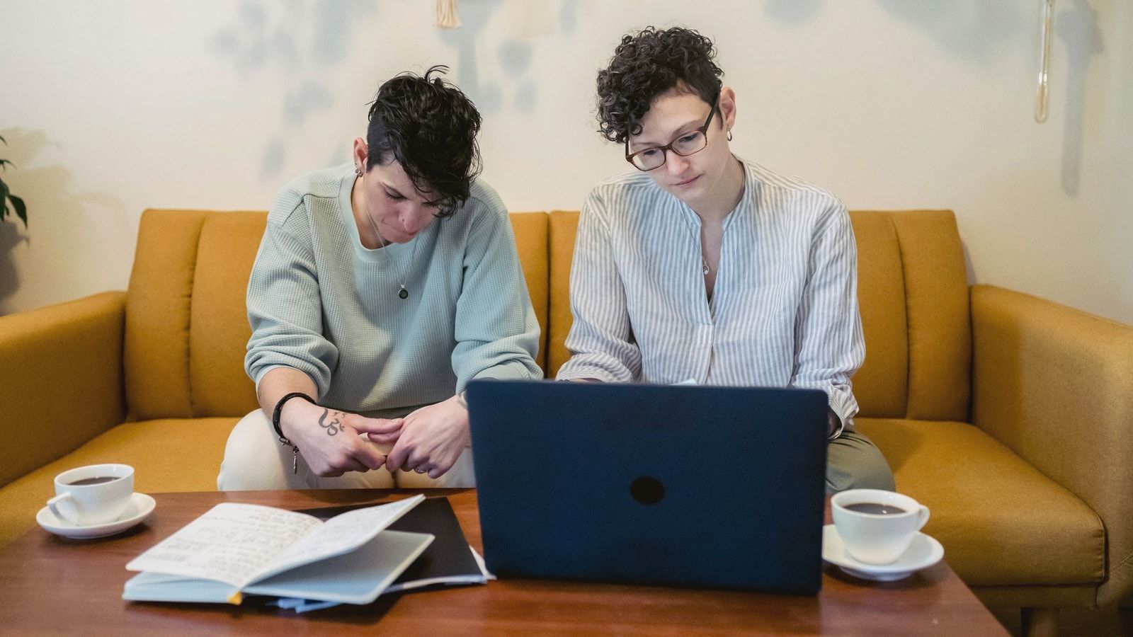 Concentrated young woman with dark short hair reading book while sitting on sofa near girlfriend working remotely on laptop at home