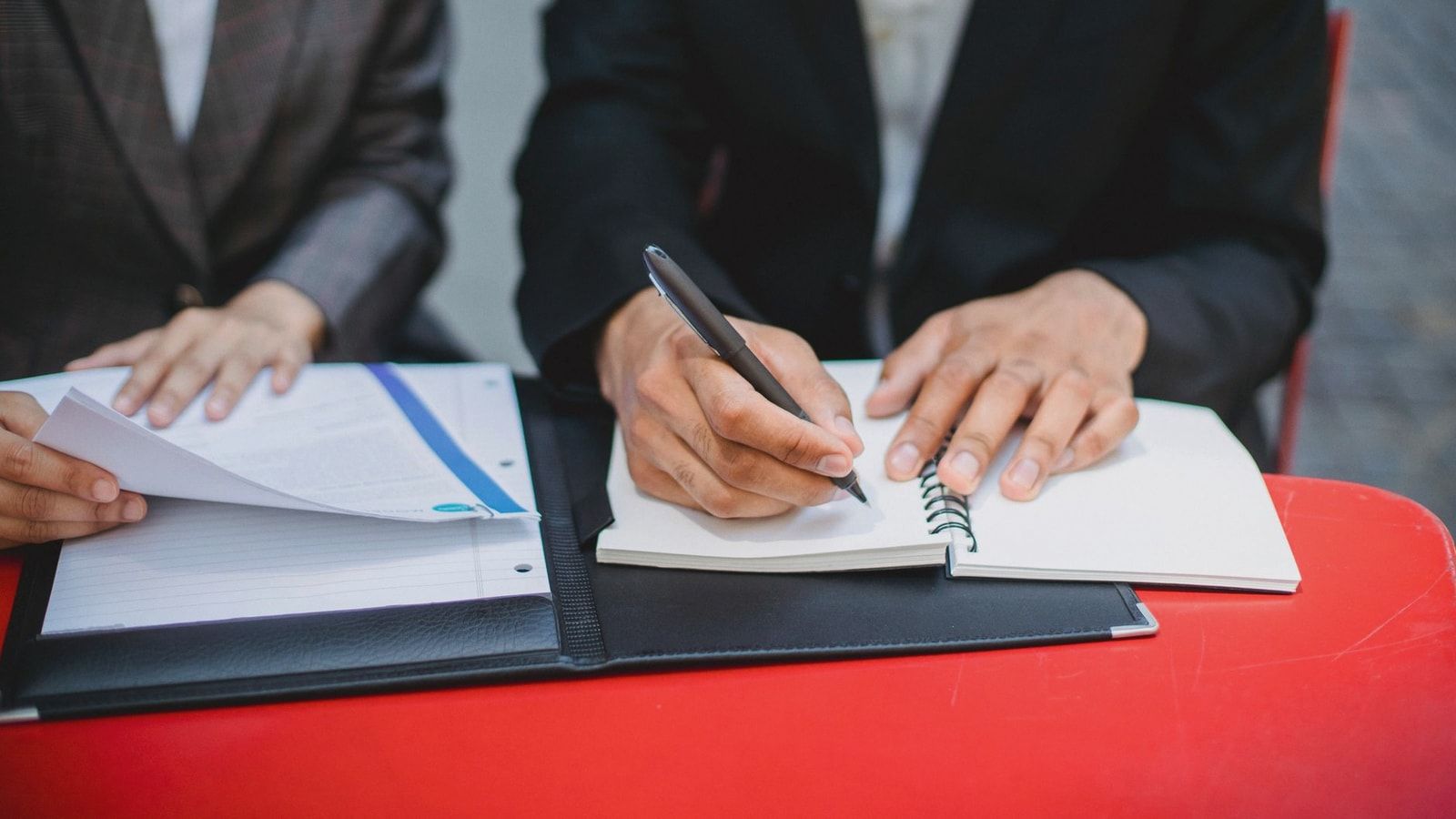 Two professionals in suits writing notes and reviewing documents at an outdoor meeting table.