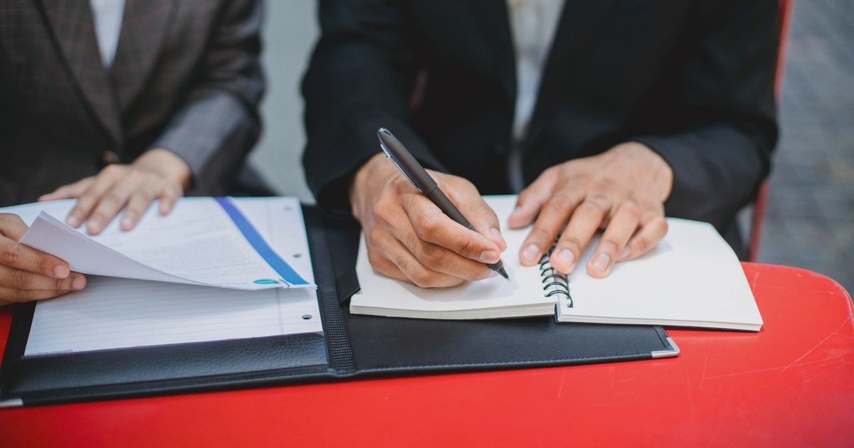 Two professionals in suits writing notes and reviewing documents at an outdoor meeting table.